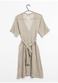 Beige stickad omslagklänning med korta ärmar, V-ringning och ett tygband i midjan. Mjuk textur och avslappnad design. Hänger från en svart krok.