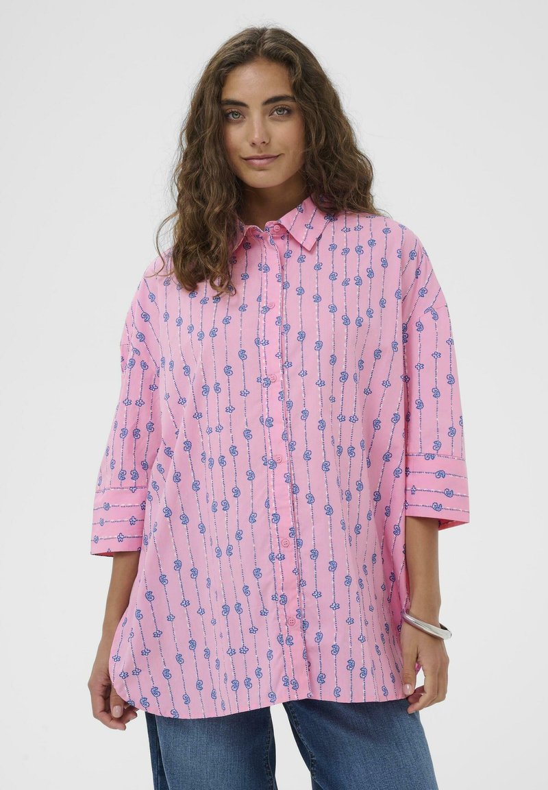 Kobieta w luźnej różowej koszuli na guziki z niebieskim kwiatowym wzorem i rękawami do łokcia, w połączeniu z niebieskimi dżinsami i srebrną bransoletką.