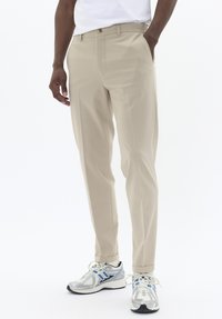 Pantalon beige au design droit, avec des poches latérales, une fermeture à bouton et une texture lisse, assorti à des baskets argentées et bleues.