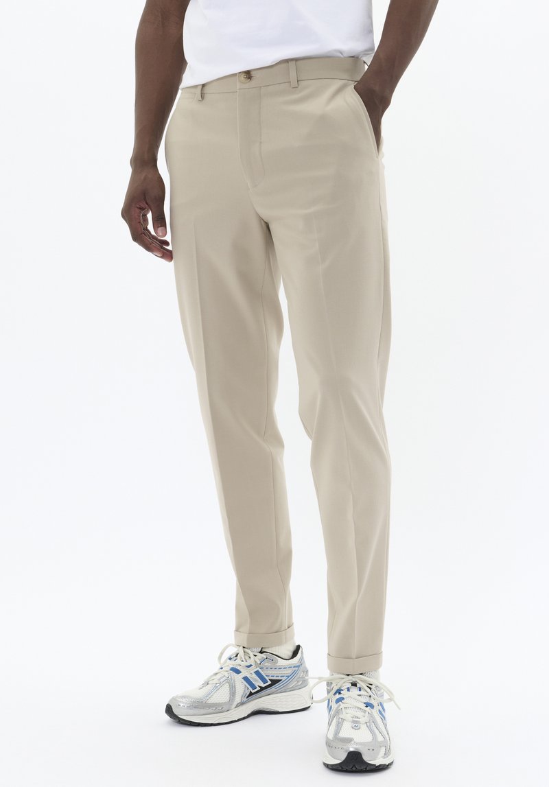 Pantalon beige au design droit, avec des poches latérales, une fermeture à bouton et une texture lisse, assorti à des baskets argentées et bleues.