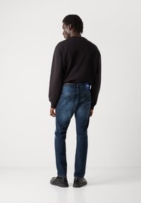 Jeans oscuros ajustados con un desvanecimiento sutil, combinados con un sudadera negra. Presenta una etiqueta de la marca azul en la cinturilla.