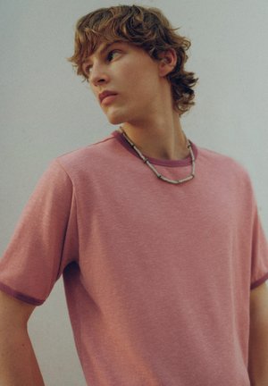Jeune personne aux cheveux courts et bouclés, portant un t-shirt rose et un collier de perles, regardant sur le côté sur un fond uni.