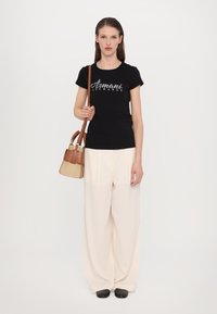 Schwarzes T-Shirt mit weißem "Armani Exchange"-Logo, beige weit geschnittene Hose und eine kleine braune Handtasche mit strukturierter Oberfläche. Schwarze Schuhe.