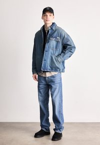 Τζάκετ από τζιν με τσέπη, ανοιχτό μπλε απόχρωση, μακριά μανίκια, κλείσιμο με κουμπιά, φορεμένο πάνω από ένα μαύρο φούτερ με φερμουάρ και κρεμ πουκάμισο, συνδυασμένο με μπλε τζιν και μαύρα παπούτσια.
