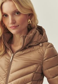 Doudoune marron clair avec une fermeture éclair et une capuche, présentant un design matelassé, une texture lisse et des boutons ronds en guise d'accent.