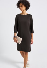 Svart klänning med trekvartslånga ärmar, rund halsringning, sidöppningar, i kombination med en vävd beige clutch och vita sneakers. Slät tygtextur.