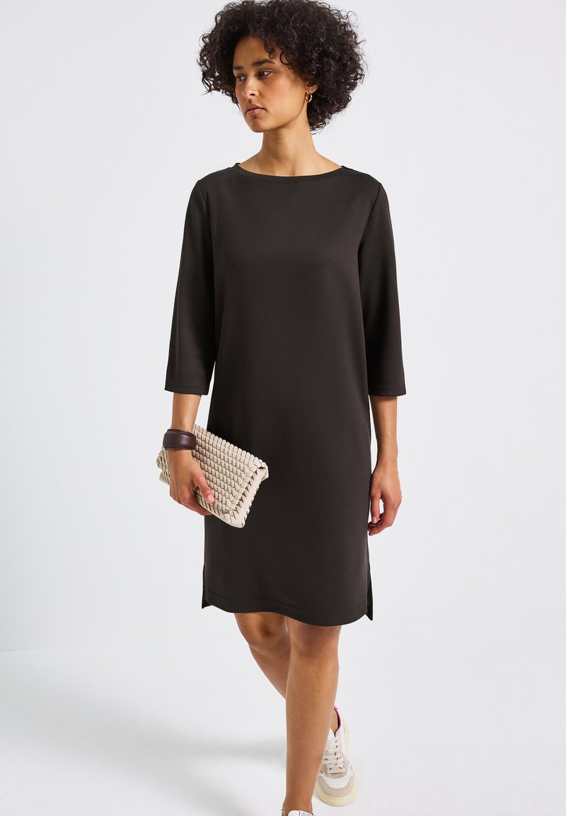 Svart klänning med trekvartslånga ärmar, rund halsringning, sidöppningar, i kombination med en vävd beige clutch och vita sneakers. Slät tygtextur.