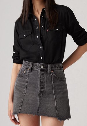 Femme portant une chemise noire boutonnée avec les manches retroussées et une mini-jupe en jean grise taille haute à ourlet effiloché.