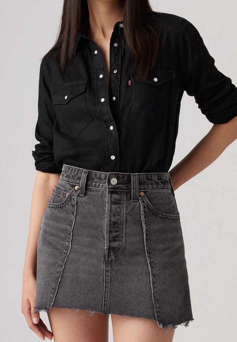 Femme portant une chemise noire boutonnée avec les manches retroussées et une mini-jupe en jean grise taille haute à ourlet effiloché.
