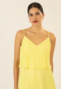 Ärmelloses, gelbes, plissiertes Kleid mit V-Ausschnitt. Verfügt über dünne Träger und dezente Perlenakzente. Leichtes Material, fließende Silhouette.