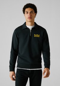 Zwarte rits-sweatshirt met hoge kraag, voorzien van "Red Bull RACING" geborduurd in het geel op de borst. Zacht stof met geribbeld manchetten.