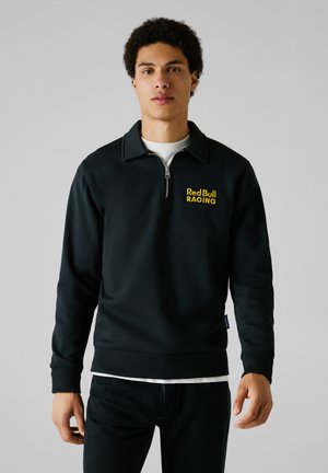 Black zip-up sweatshirt with high collar, featuring "Red Bull RACING" embroidered in yellow on the chest. Soft fabric with ribbed cuffs.