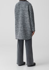 Abrigo texturizado en gris y negro con un collar plano, mangas largas y un dobladillo ligeramente curvado, combinado con pantalones grises de campana y zapatos planos.