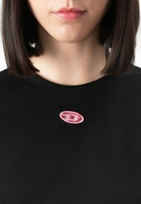 Close-up of a person wearing a black shirt with a small pink oval-shaped embroidered logo near the collar.