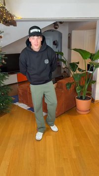 Μαύρο φούτερ με κουκούλα και λογότυπο, χακί παντελόνι και λευκά αθλητικά παπούτσια. Εσωτερικός χώρος με ξύλινο πάτωμα και γλάστρα κοντά.