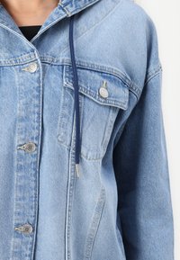 Svetlo modra denim jakna s kapuco, srebrnimi pritisnimi gumbi, mornarsko modrim veznim trakom, žepom na prsih in detajli šivanja ob šivih.