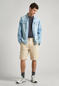 Giacca di jeans azzurro chiaro con due tasche sul petto, indossata sopra un top scuro, abbinata a pantaloni beige e sneakers bianche con calzini grigi.