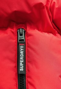Sarkanais pūkainais jaka audums ar spīdīgu apdari. Melna rāvējslēdzējs ar balto "SUPERDRY" logo un šūšanas detaļām šuvē.