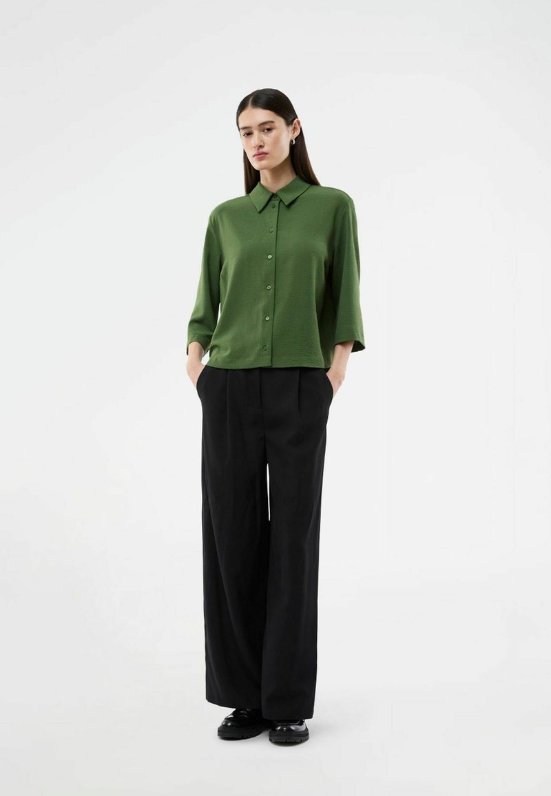 Femme debout les mains dans les poches, portant une chemise verte boutonnée et un pantalon noir à jambes larges sur un fond blanc uni.