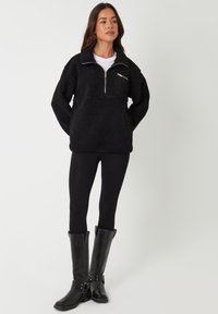 Sort fleece trøje med lynlås halvvejs og forlomme, parret med sorte leggings og knæhøje sorte læderstøvler.