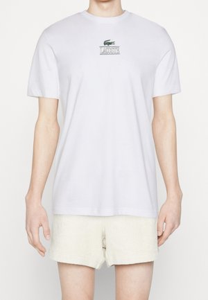 Homme portant un t-shirt blanc uni à manches courtes avec un petit logo Lacoste vert et un short texturé couleur crème, debout devant un fond blanc.