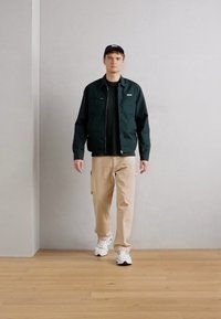 Donkergroene zip-up jack over een zwart shirt, beige cargobroek en witte sneakers. Het model staat op een lichte houten vloer tegen een grijze muur.