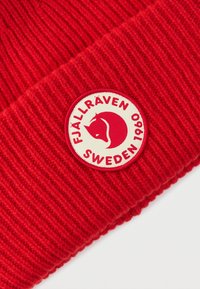 Κόκκινος ριμπ πλεκτός σκούφος με κυκλικό κρεμ μπατς που φέρει τη φράση "FJÄLLRÄVEN SWEDEN 1960" και ένα λογότυπο με αλεπού σε κόκκινο. Μαλακή υφή από μάλλινη ύλη.