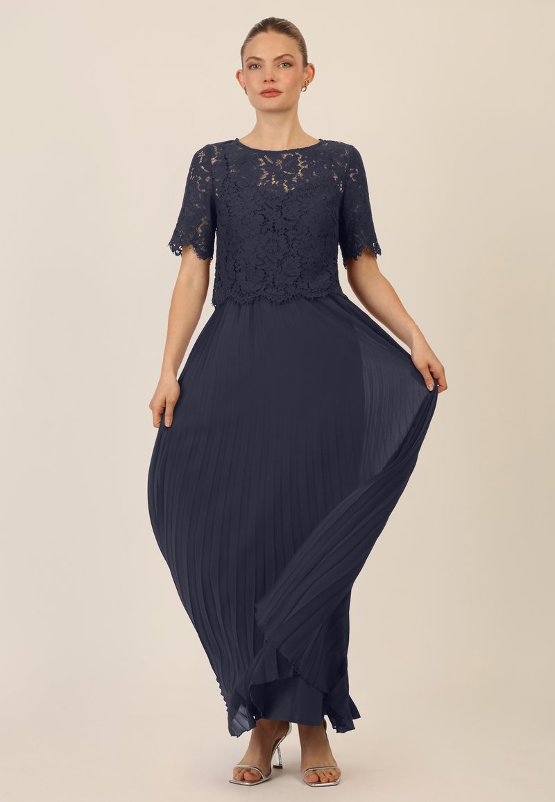 Navyblaues Kleid mit Spitzenoberteil und kurzen Ärmeln, das mit einem gefältelten Rock, der elegant fließt, ergänzt wird. Dazu passende silberne Absätze.