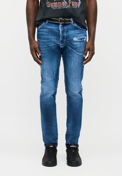 Homme portant un jean slim bleu délavé avec légère usure, ceinture noire cloutée, baskets noires et un T-shirt noir "Summer Pump".