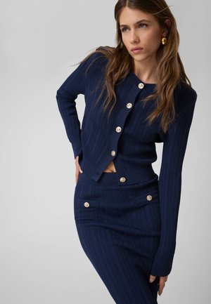 Femme portant un cardigan en maille côtelée bleu marine avec des boutons dorés et une jupe assortie, posant avec une main sur la hanche sur un fond gris.