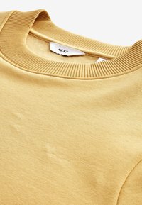 Sweat-shirt jaune moutarde clair avec encolure ronde côtelée et étiquette de marque blanche visible à l'intérieur du col.