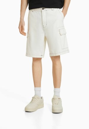 Pantalones cortos cargo blancos con costuras en contraste, de longitud hasta la rodilla, que cuentan con bolsillos laterales y cierre de botón. Se usan con zapatillas y calcetines de color claro.
