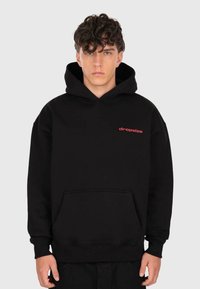 Junger Mann mit lockigem Haar, der einen schwarzen Hoodie mit Fronttasche und kleinem roten "dropsize"-Text auf der linken Brust trägt.