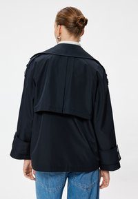 Giacca trench nera oversize con retro a strati, maniche ampie e polsini abbottonati, realizzata in tessuto morbido, abbinata a jeans blu chiaro.