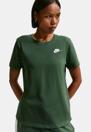 Donna con una T-shirt Nike verde scuro con logo bianco, che guarda di lato, con le mani dietro la schiena, su uno sfondo chiaro uniforme.