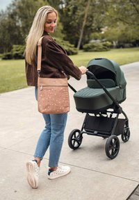 Tan crossbody taška s květinovým vzorem, hnědá manšestrová bunda, modré džíny a bílé tenisky, tlačící zelený kočárek po cestě.