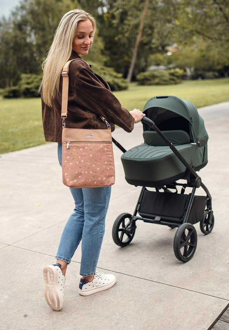 Tan crossbody taška s květinovým vzorem, hnědá manšestrová bunda, modré džíny a bílé tenisky, tlačící zelený kočárek po cestě.