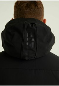 Persona con cabello corto que lleva una chaqueta negra con capucha levantada que muestra el texto vertical "NASSIN" en la parte trasera.