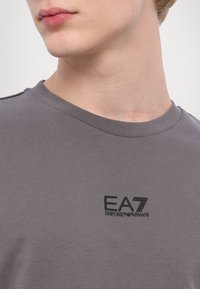 Szürke pamut póló, kerek nyakkal és rövid ujjal. Az mellkason fekete EA7 Emporio Armani logó található. Sima tapintású.