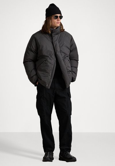 Person med sort beanie, mørke solbriller, gråt puffet jakke, sorte cargo-bukser og sorte sneakers, står med hænderne i lommerne.
