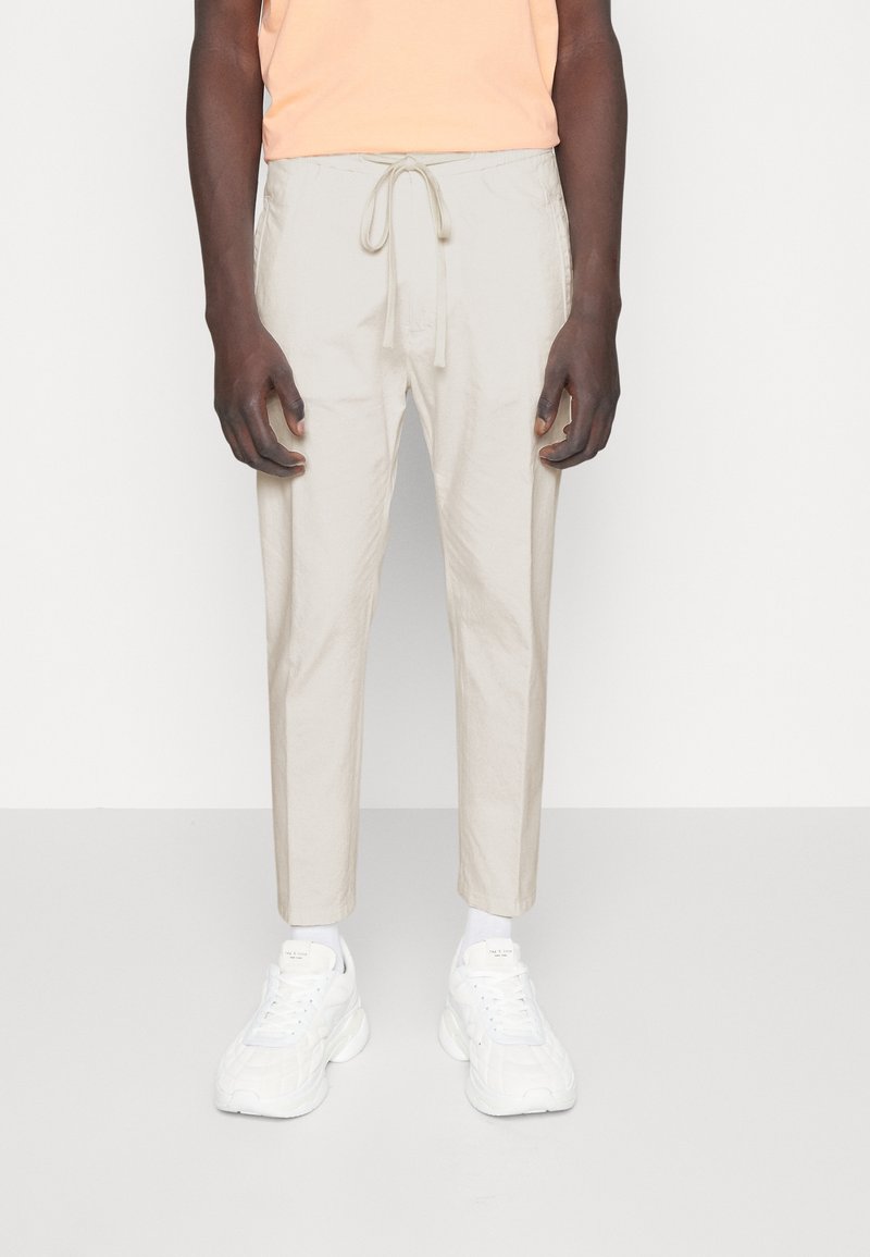 Person i beige dragsko byxor, vita strumpor och vita sneakers, stående mot en enkel ljus bakgrund.