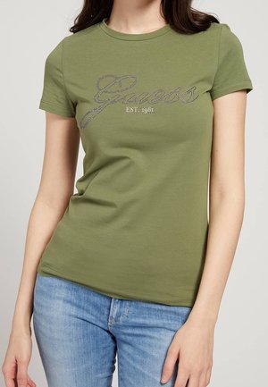 Femme portant un T-shirt ajusté à manches courtes couleur vert olive avec le logo "Guess Est. 1981" en strass, associé à un jean bleu clair.
