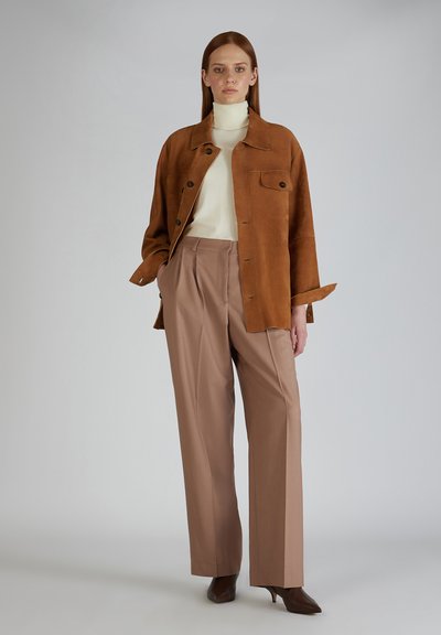 Bruine suède jas met twee borstzakken, crèmekleurige coltrui en beige high-waisted broek, aangevuld met bruine hakken.