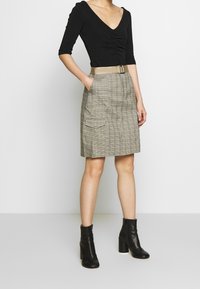 Haut noir à col en V avec des détails froncés ; jupe à carreaux beige avec poches et ceinture assortie ; bottines à talons noires.