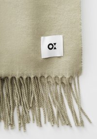 Zachte beige wollen sjaal met gedraaide franjes en een klein wit rechthoekig label met daarop een zwart geometrisch logo.