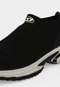 Μαύρο αθλητικό παπούτσι slip-on με λευκή σόλα και υφασμάτινο, ριγωτό επάνω μέρος που δείχνει ένα μικρό οβάλ λογότυπο κοντά στον αστράγαλο.