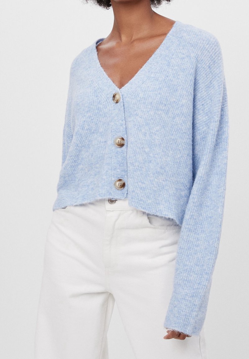 Kvinna iförd en ljusblå knappad kofta med V-ringning och vita högmidjade byxor, visad från halsen till höfterna mot en enfärgad bakgrund.