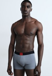 Szürke boxer alsónadrág fekete derékpánttal, melyen egy kis piros logó található. Az anyag simának és testhez simulónak tűnik, hangsúlyozva a viselő fizikáját.