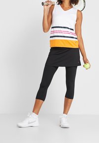 Top bianco senza maniche con righe orizzontali multicolori e orlo arancione, abbinato a uno skort nero. La modella tiene una racchetta da tennis e una pallina.