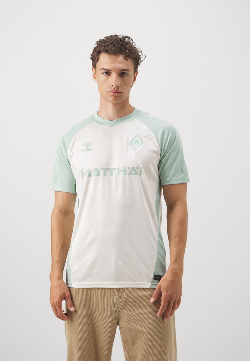 Maglia sportiva bianca con accenti verde menta, caratterizzata da un colletto a girocollo, maniche corte, motivo a rombi e logo frontale. Indossata con pantaloni in velluto a coste beige.